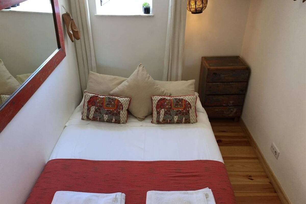 Una camera da letto con un letto, cuscini, una coperta rossa, due asciugamani piegati, una cassettiera in legno, uno specchio e una finestra con tende.