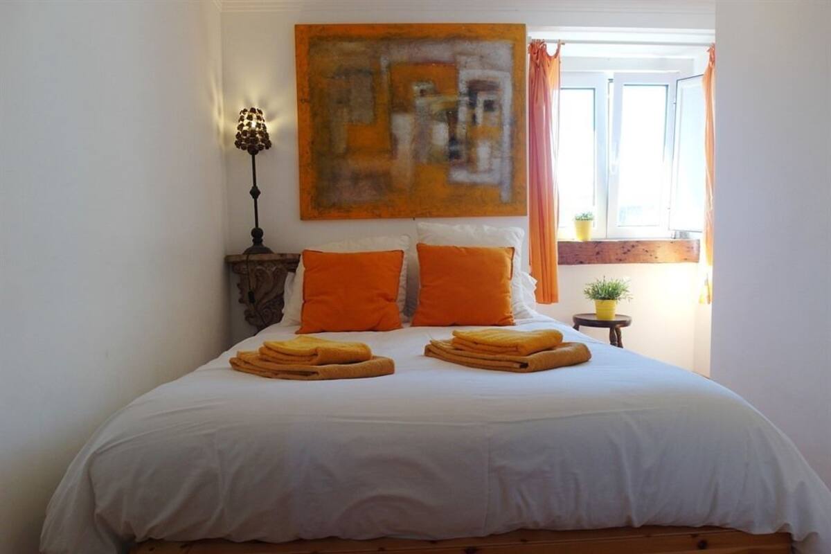 Un letto rifatto con cura, con lenzuola bianche e arancioni, una lampada, un quadro e una finestra con tende.