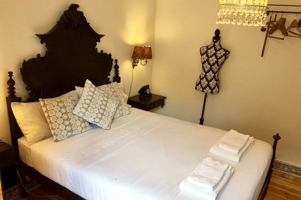 Un letto rifatto con lenzuola bianche e cuscini a fantasia, una testiera in legno, un comodino con una lampada e un manichino per abiti.