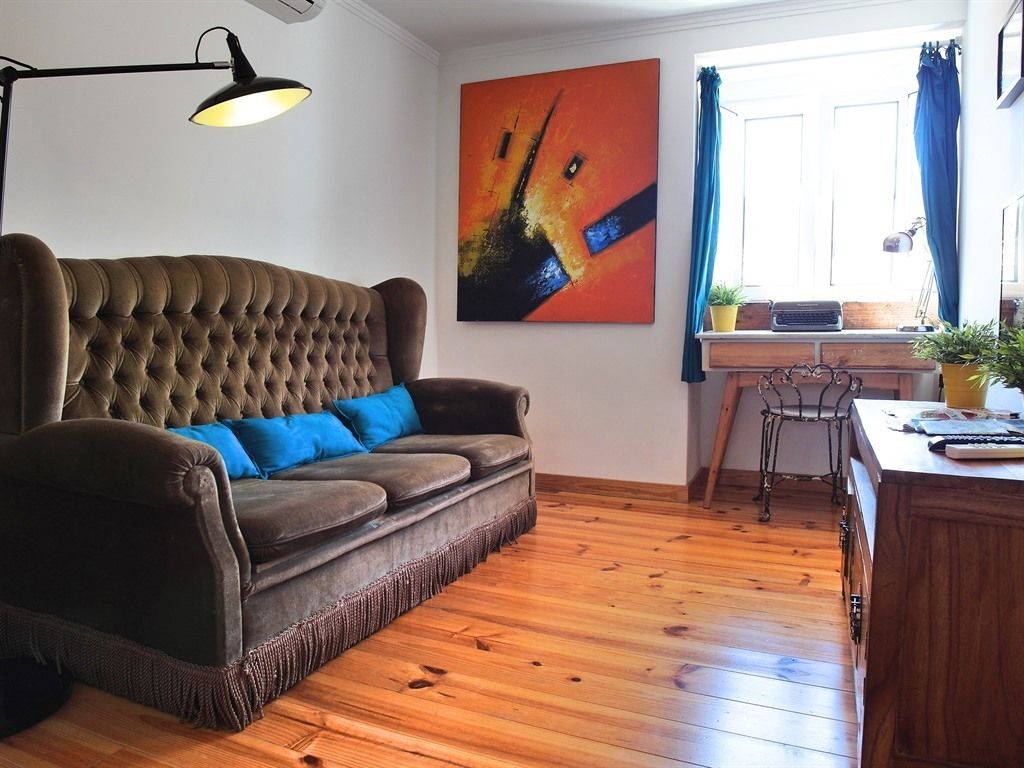 Ein Wohnzimmer mit einem раpfen Sofa, einem Schreibtisch mit Schreibmaschine, einem Fenster mit blauen Vorhängen und einem farbenfrohen abstrakten Gemälde an der Wand.