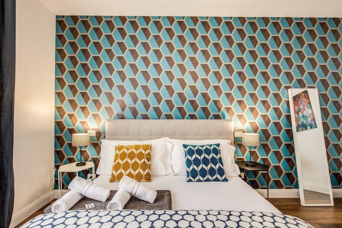 A hotel room with a geometric patterned wall, a bed with white and patterned pillows, two bedside tables with lamps, and a large mirror on the wall.