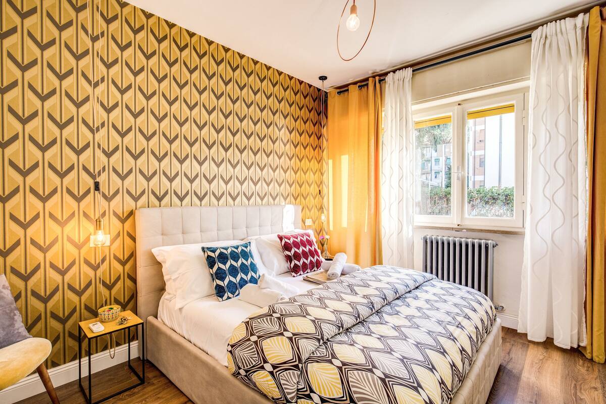 A bedroom with a patterned wallpaper, a bed with multiple pillows, a bedside table with a lamp, and a window with curtains.