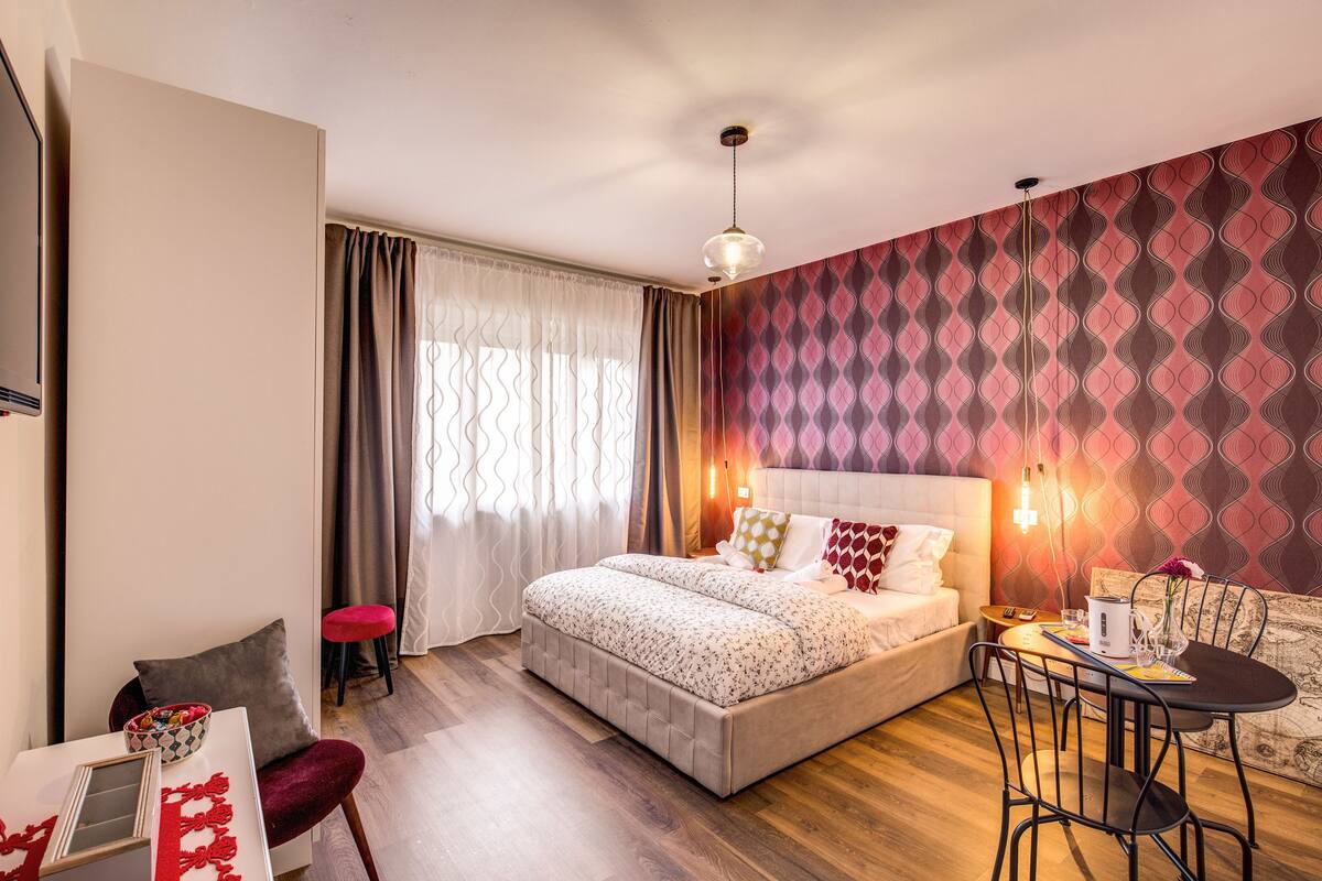 A modern hotel room with a large bed, a dining area with a table and chairs, a small stool, and a TV on a stand.