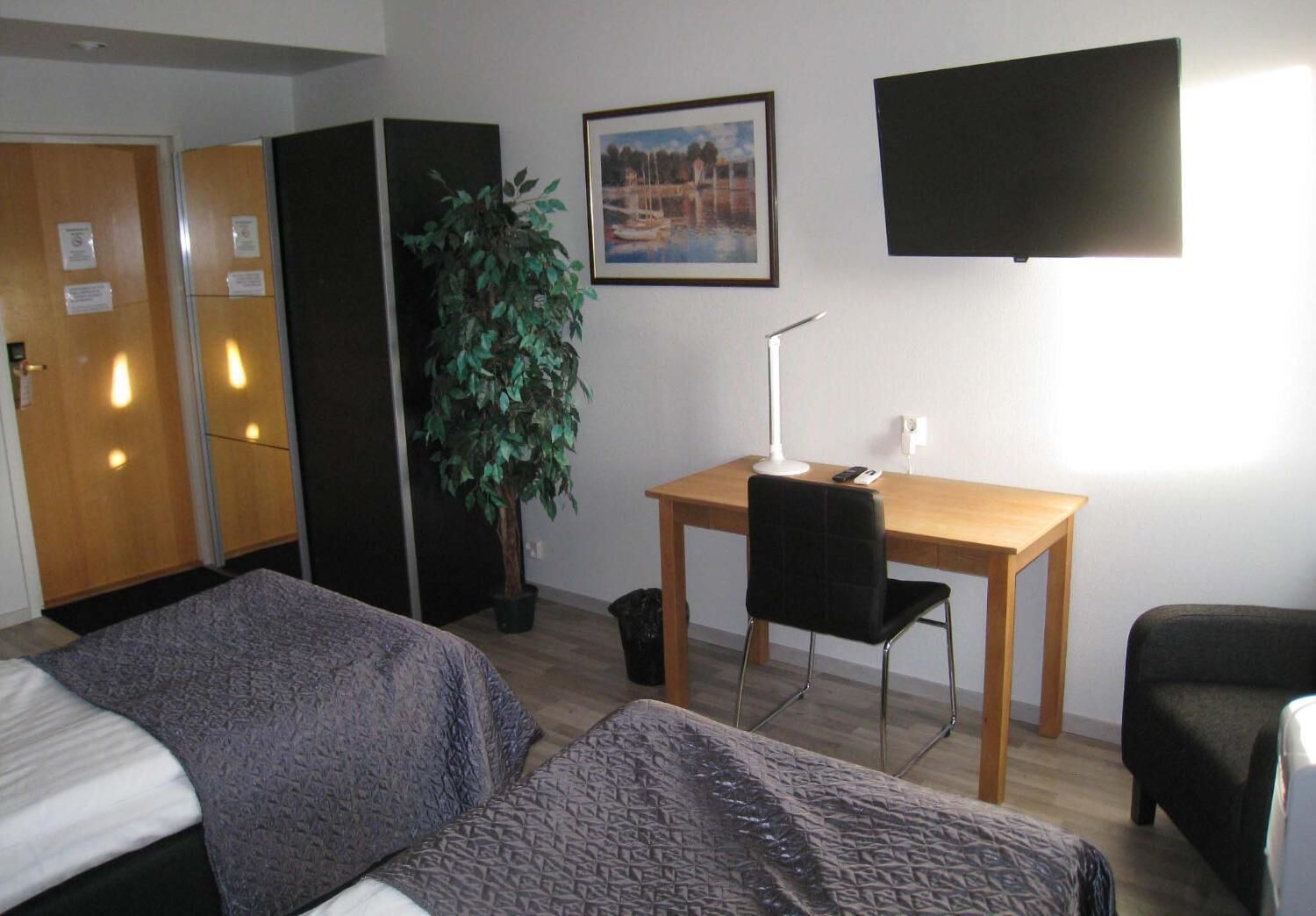 Une chambre d’hôtel avec deux lits, un bureau avec une chaise, une plante en pot, un tableau encadré au mur et un téléviseur fixé au-dessus du bureau.