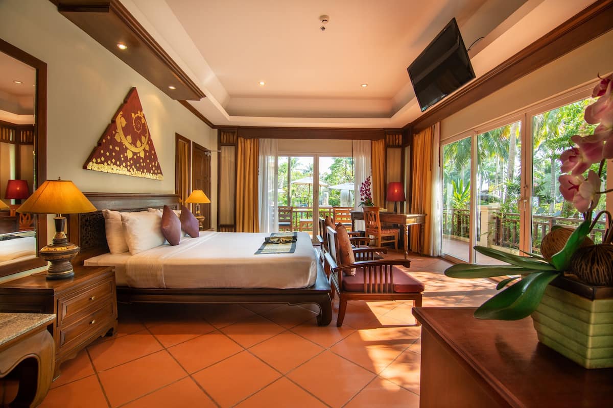 Habitación de hotel con cama, mesitas de noche, escritorio, silla, televisor y ventanas amplias con vistas a zonas verdes.