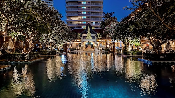 A nighttime view of a hotel with a large pool, surrounded by trees and a multi-story building in the background. - Avalon Beach Resort Pattaya (Pattaya)