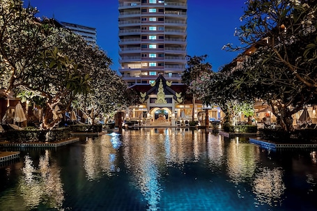 A nighttime view of a hotel with a large pool, a traditional building with a golden facade, and a tall building in the background.. Avalon Beach Resort Pattaya