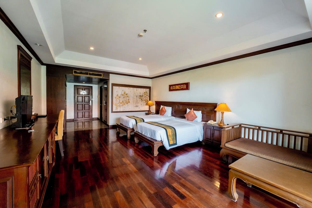 Habitación de hotel con piso de madera, una cama grande con dos almohadas, una mesita de noche con lámpara y un banco.