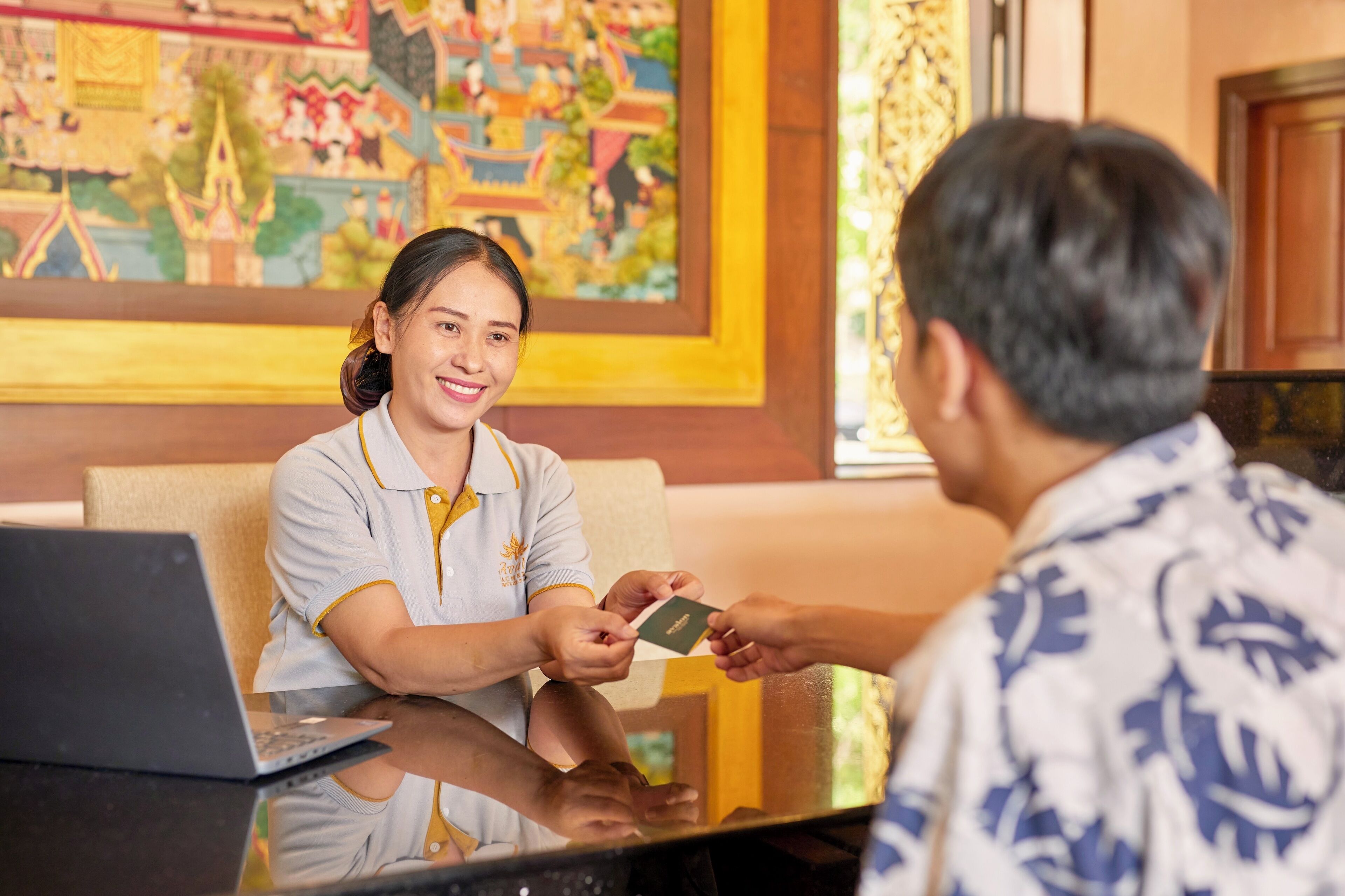 A hotel reception desk with a laptop, a person handing over a card, and a colorful mural in the background.
