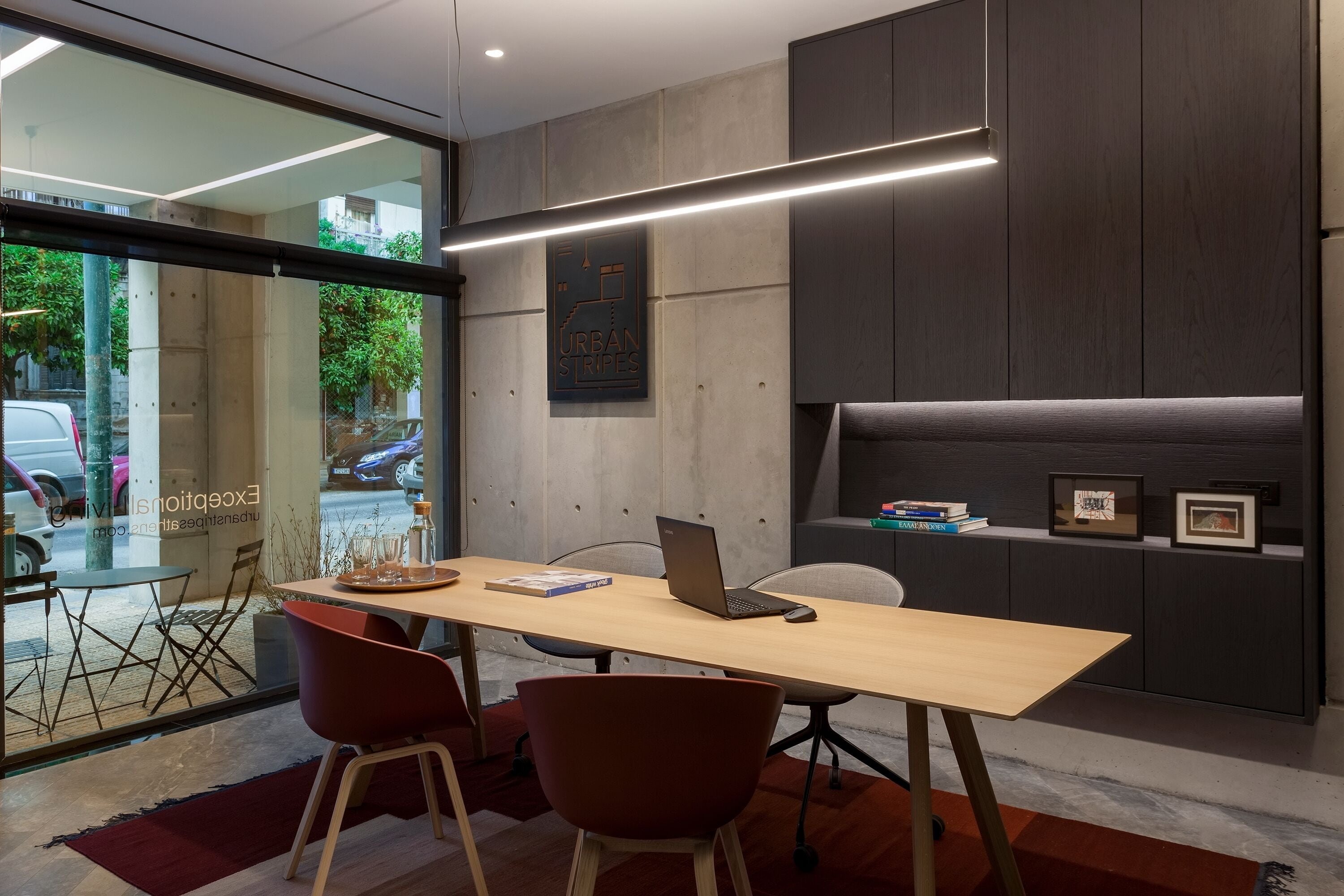 A modern office space with a wooden desk, two red chairs, a laptop, and a bookshelf with books and framed pictures.