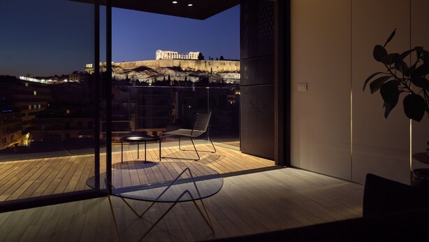 A modern balcony with a glass door, a wooden floor, a round glass table, a metal chair, and a view of a historic building at dusk. - Urban Stripes Athens (Athens)