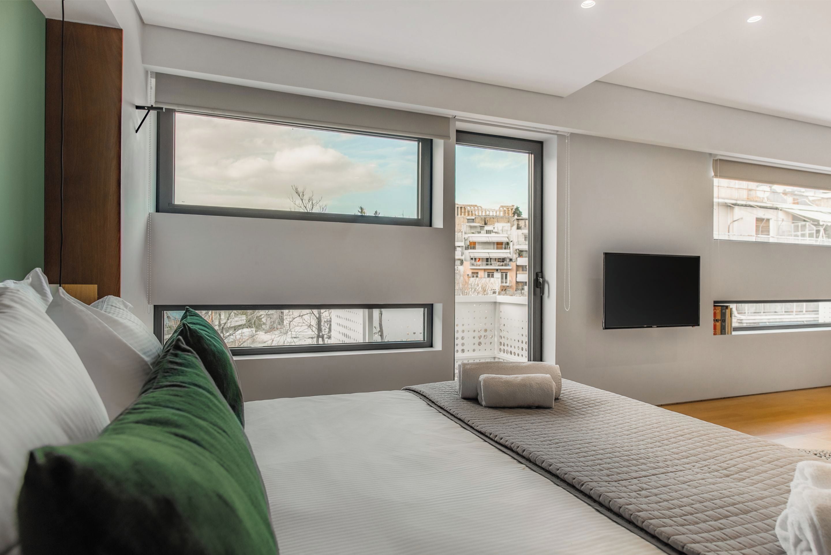 Une chambre moderne avec un grand lit, un téléviseur fixé au mur et une vue sur la ville par les fenêtres.