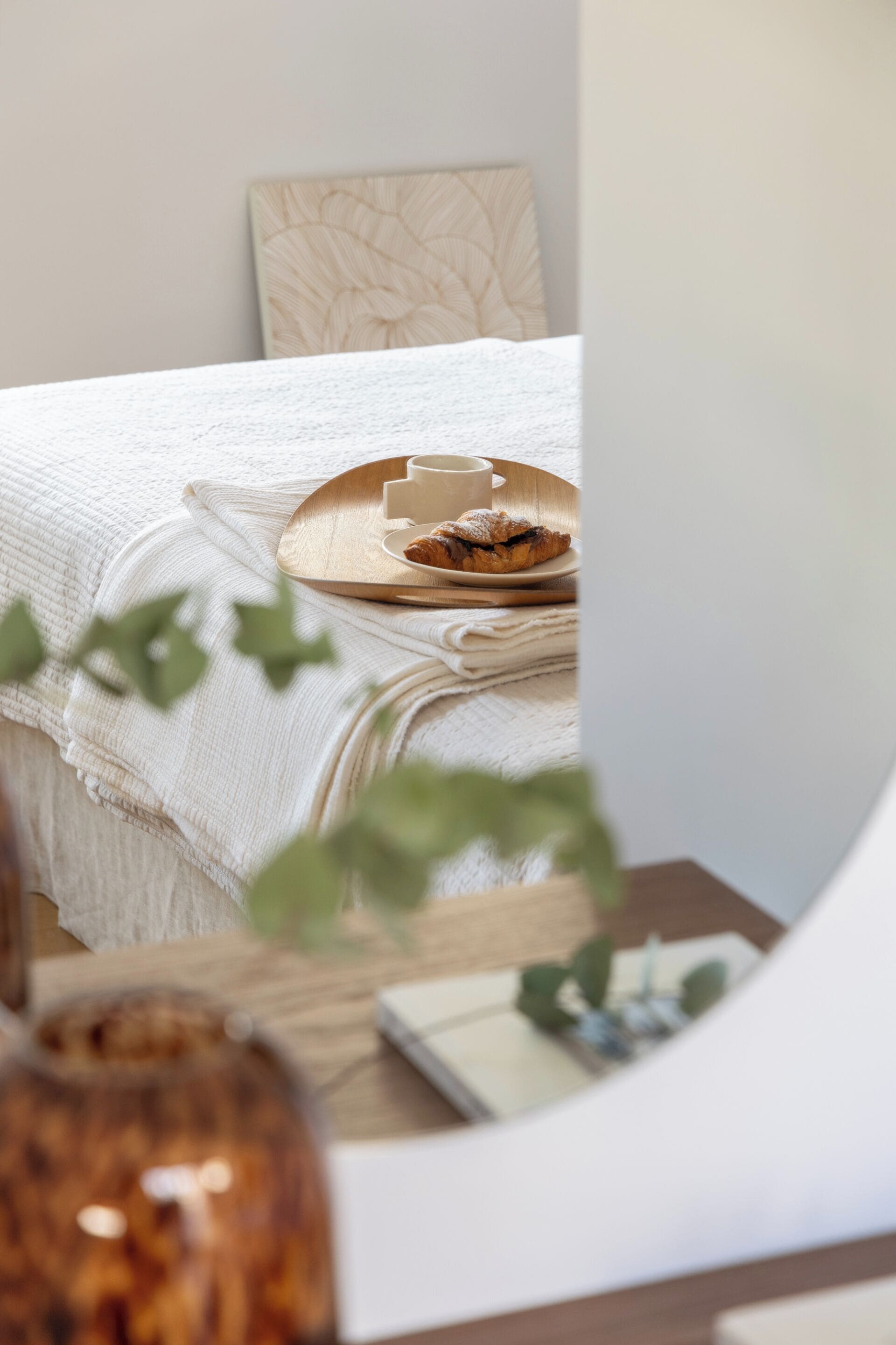 A neatly made bed with a white blanket, a wooden tray with a cup and pastry, and a plant in the foreground.