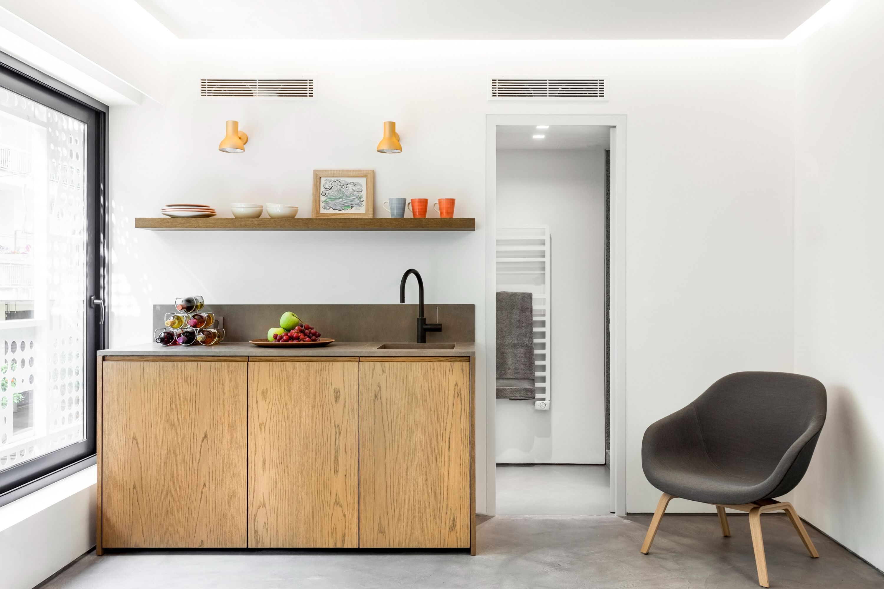A modern kitchen with wooden cabinets, a sink, and a countertop with various items. There is a chair and a door leading to another room.