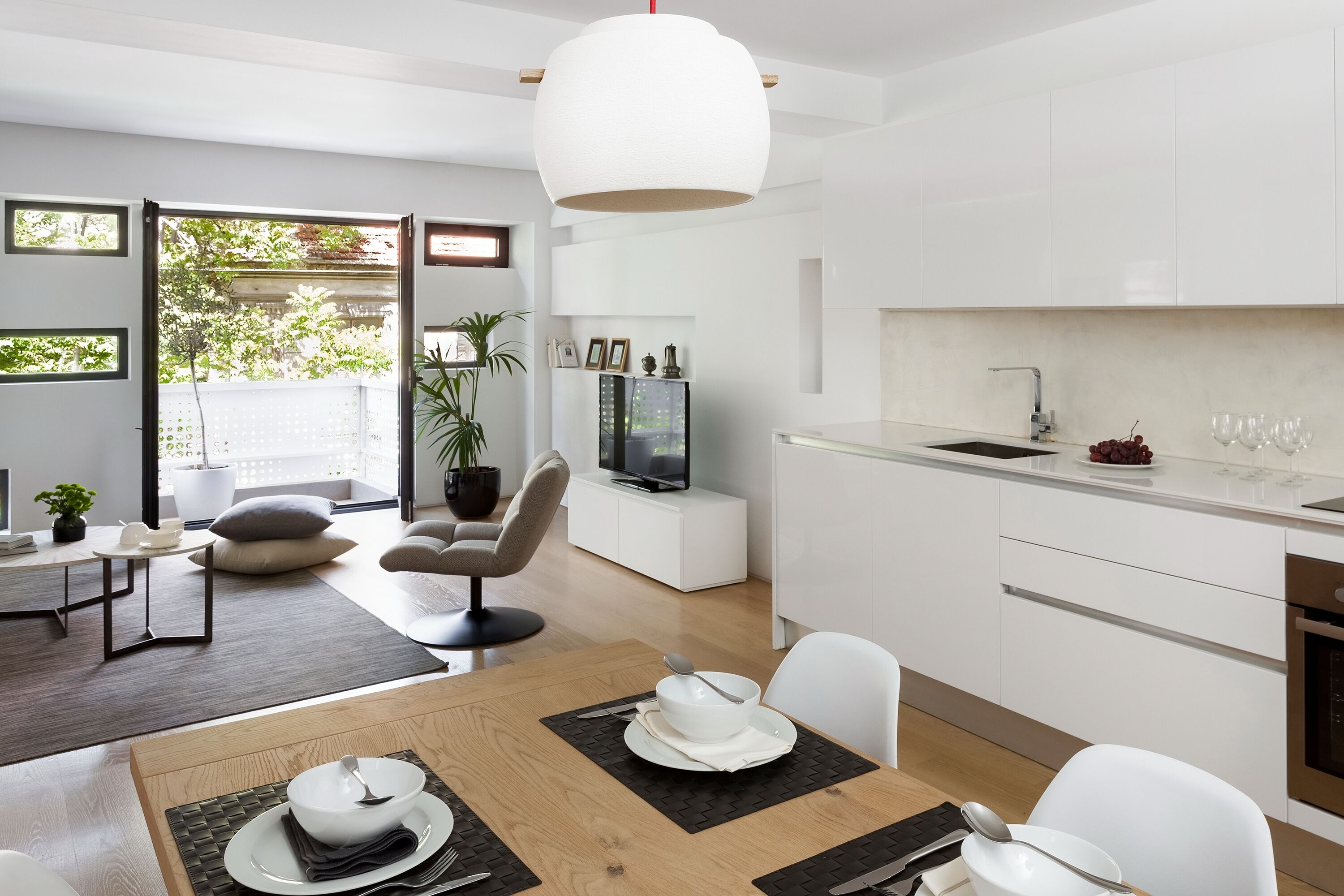 A modern kitchen with white cabinets, a dining area with a wooden table, and a TV on a white cabinet.
