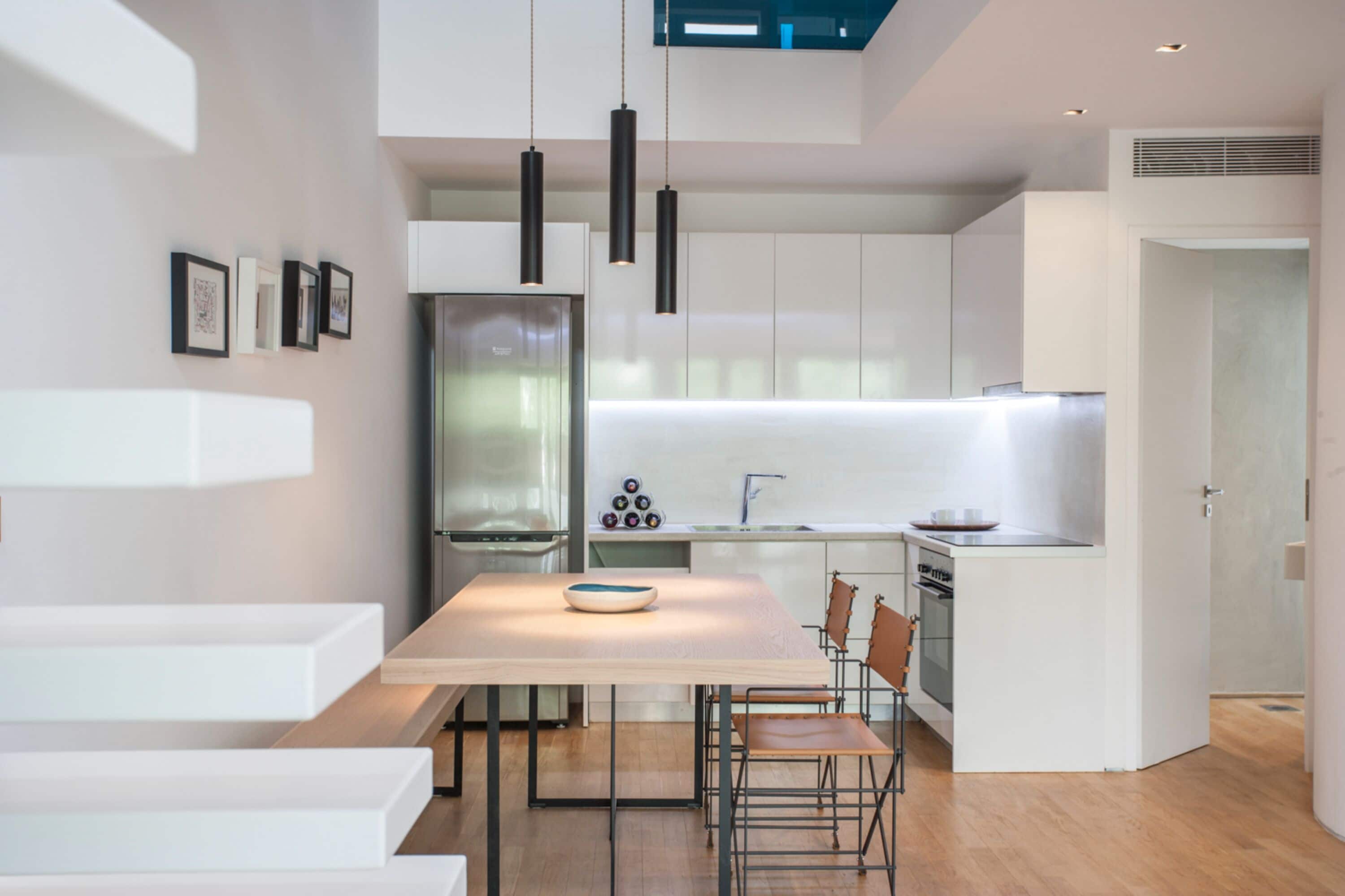 Cozinha moderna com armários brancos, uma ilha central, iluminação pendente e um espaço de jantar com cadeiras.