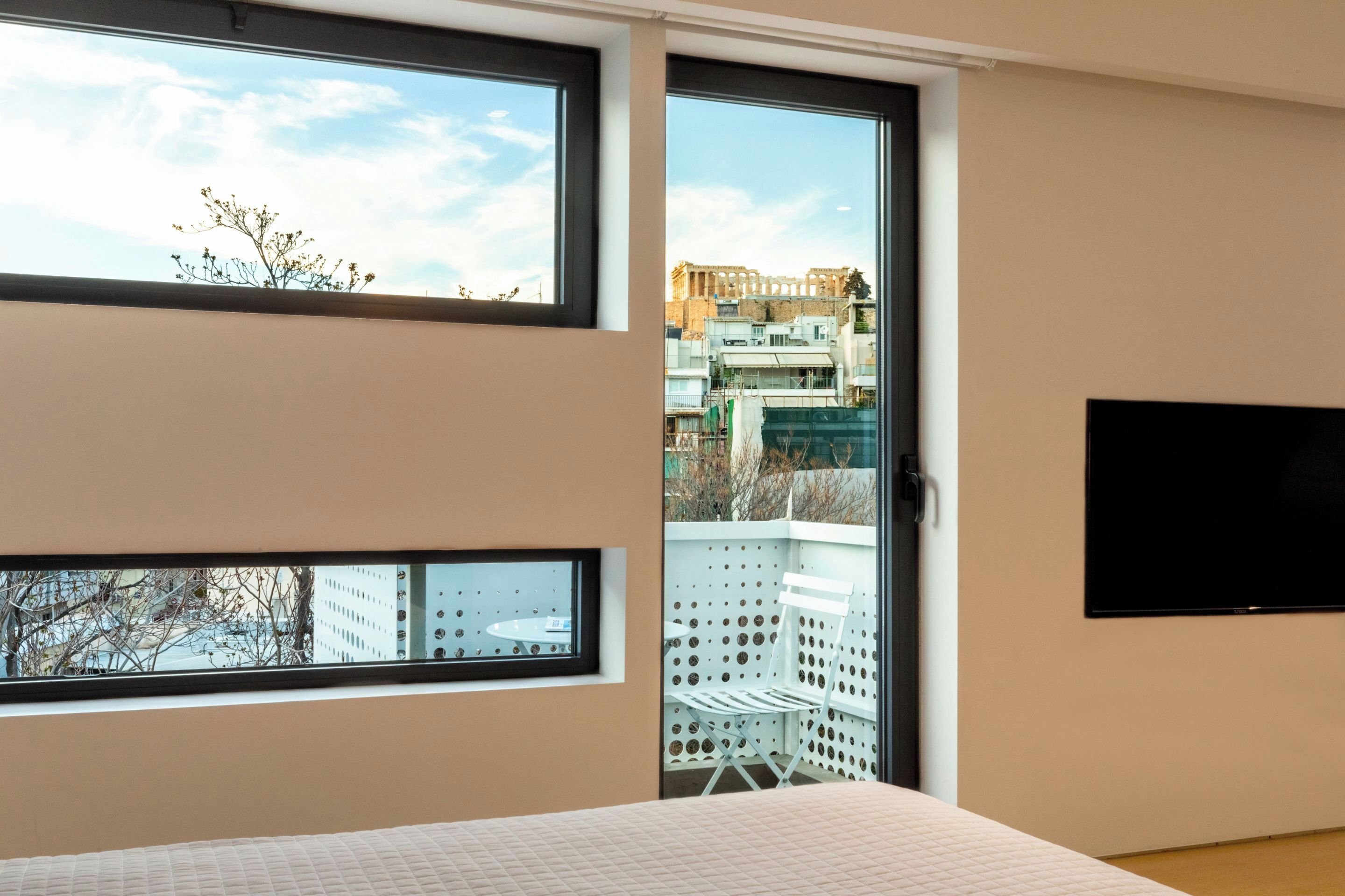 A bedroom with a large window, a bed, a TV, and a balcony with a chair.
