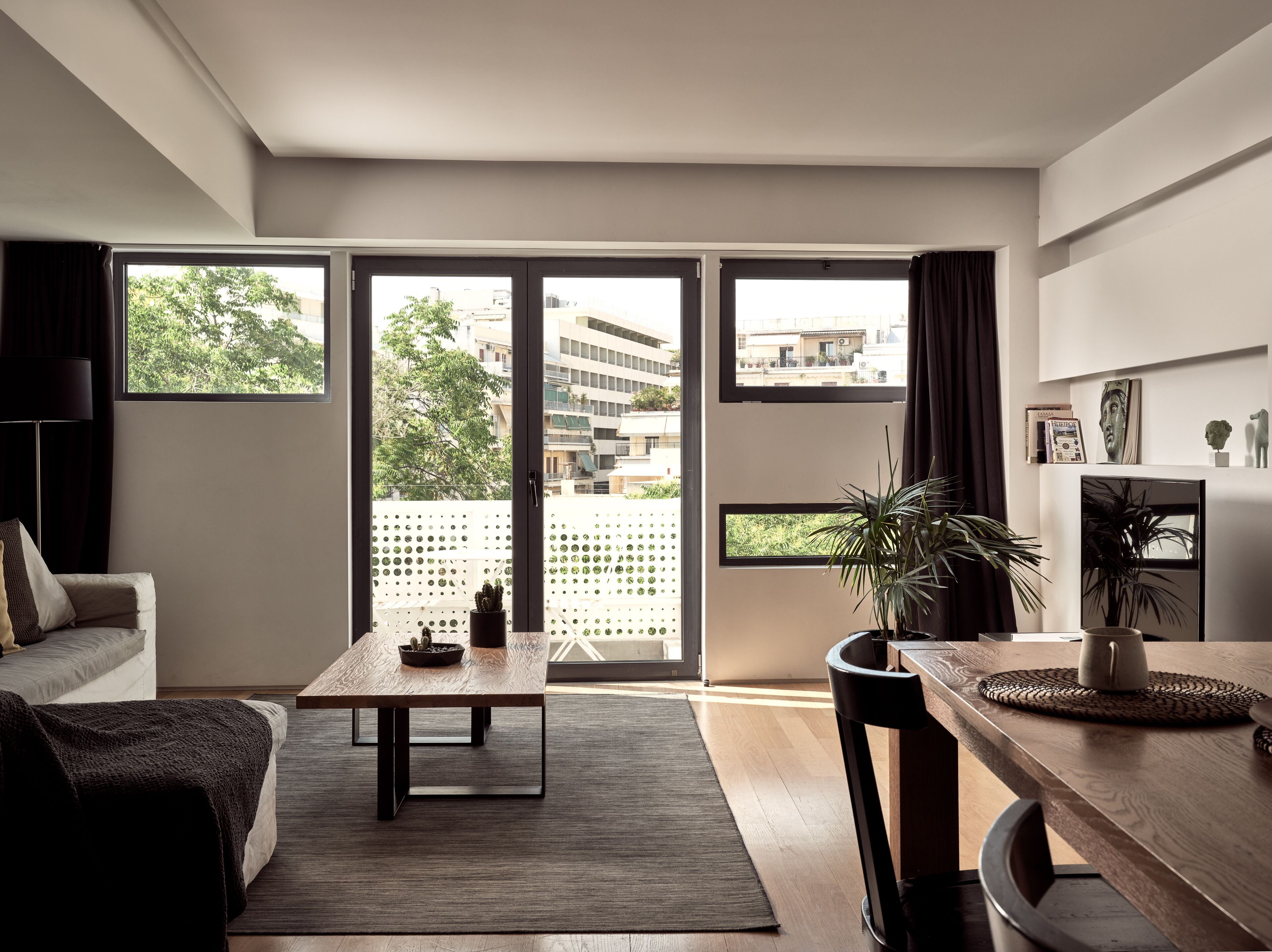 Un salon moderne avec un canapé, une table basse et un coin repas. De grandes fenêtres, ornées de rideaux, offrent une vue sur les bâtiments environnants, et une plante en pot est posée sur le rebord de la fenêtre.