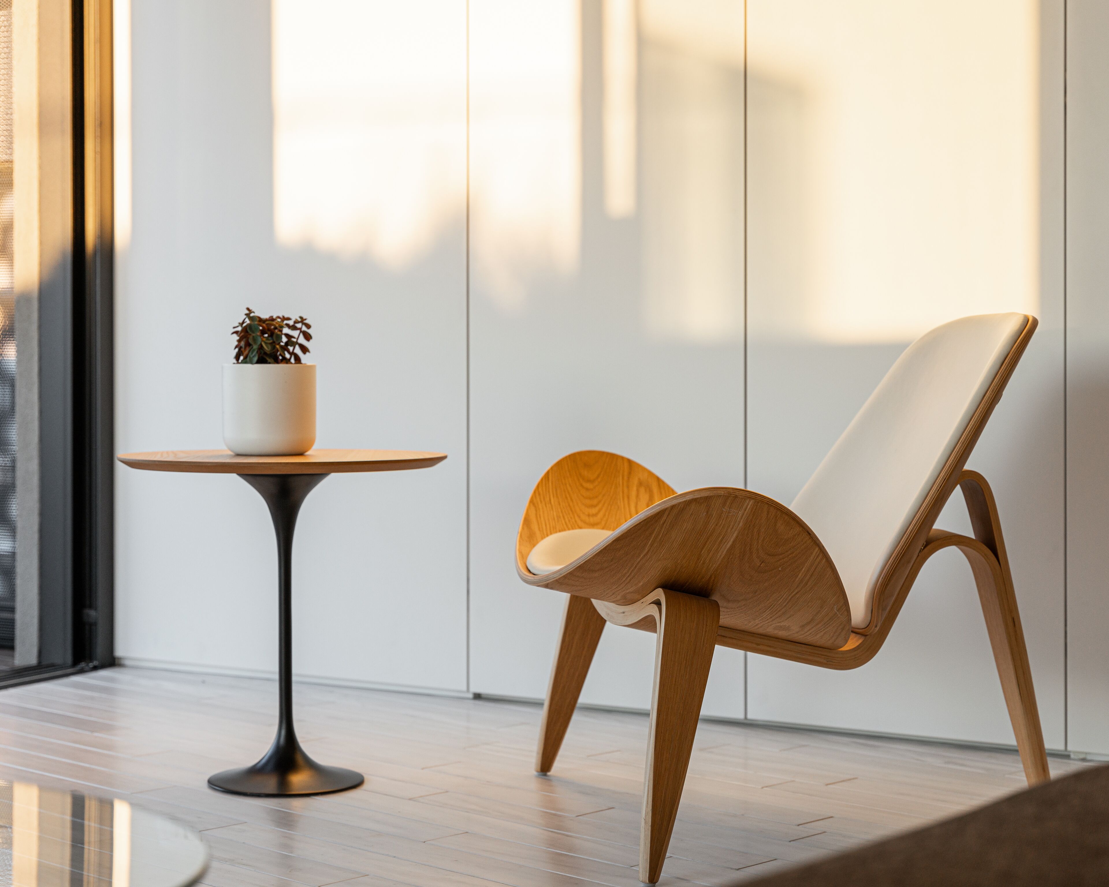 A modern room with a wooden floor, a round table with a potted plant, and a wooden chair with a white cushion.