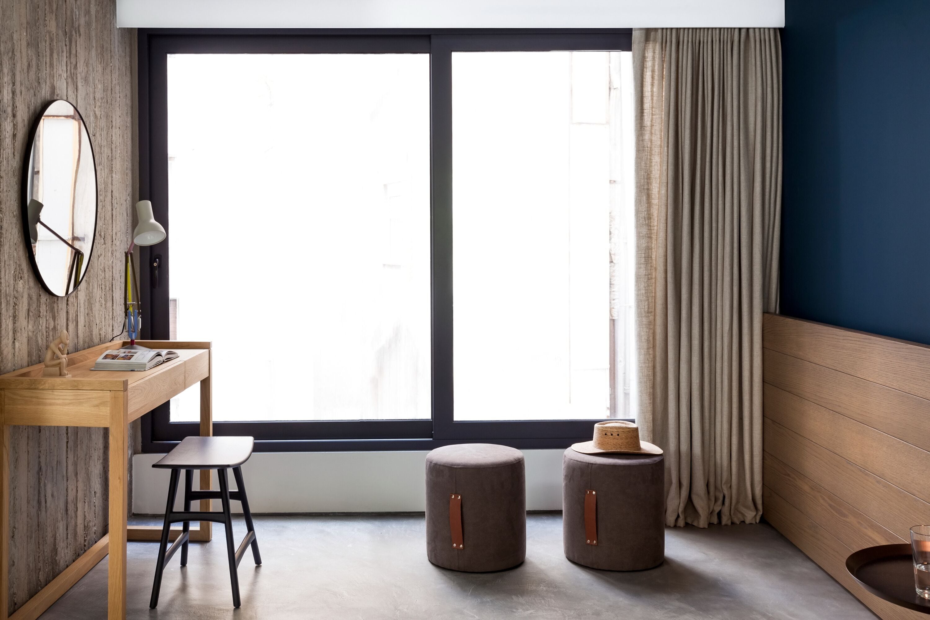 A modern room with a wooden desk, a chair, a round stool, a mirror, and a large window with curtains.