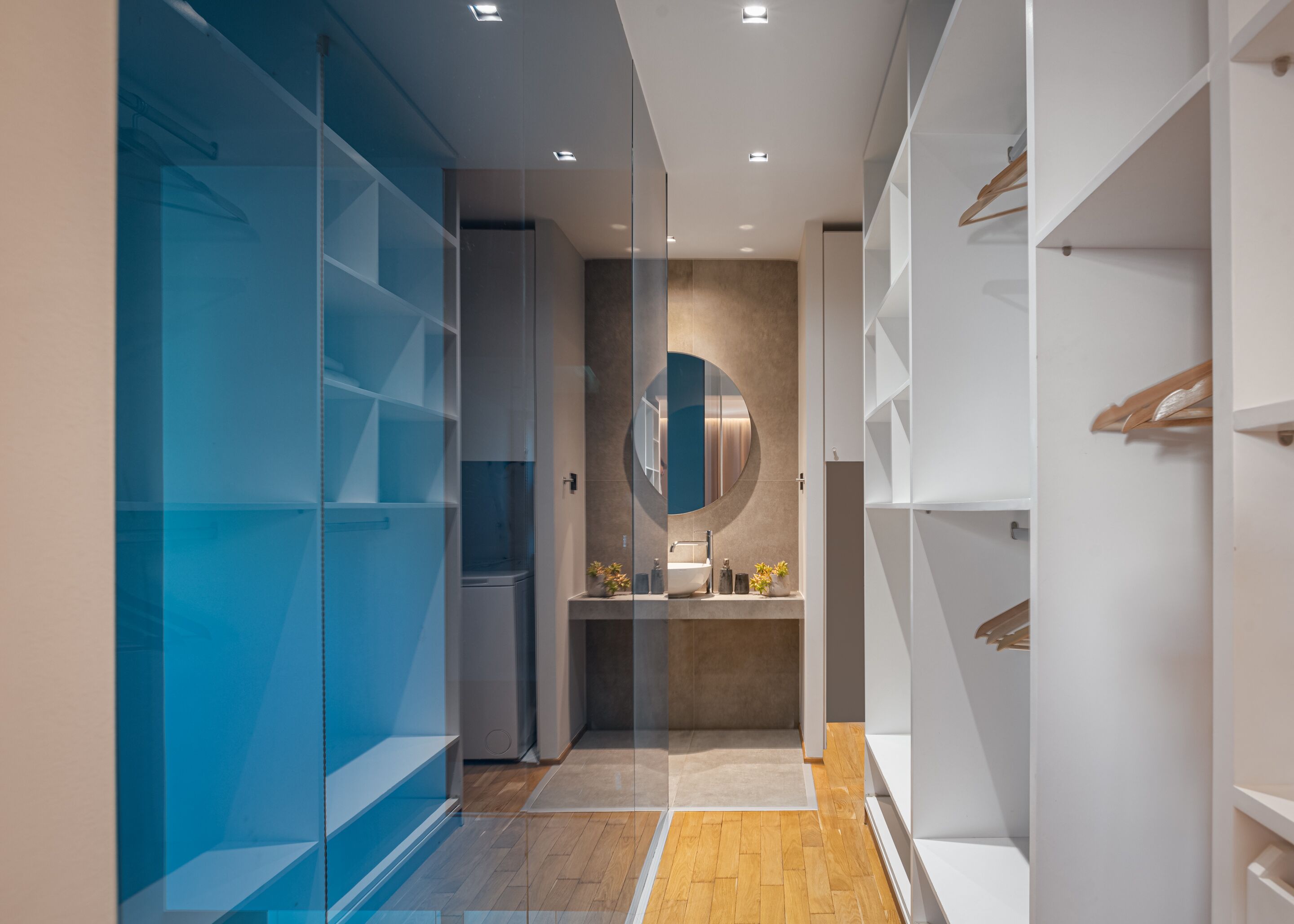 A modern bathroom with a glass partition, a wooden floor, a sink with a mirror, and white storage shelves.