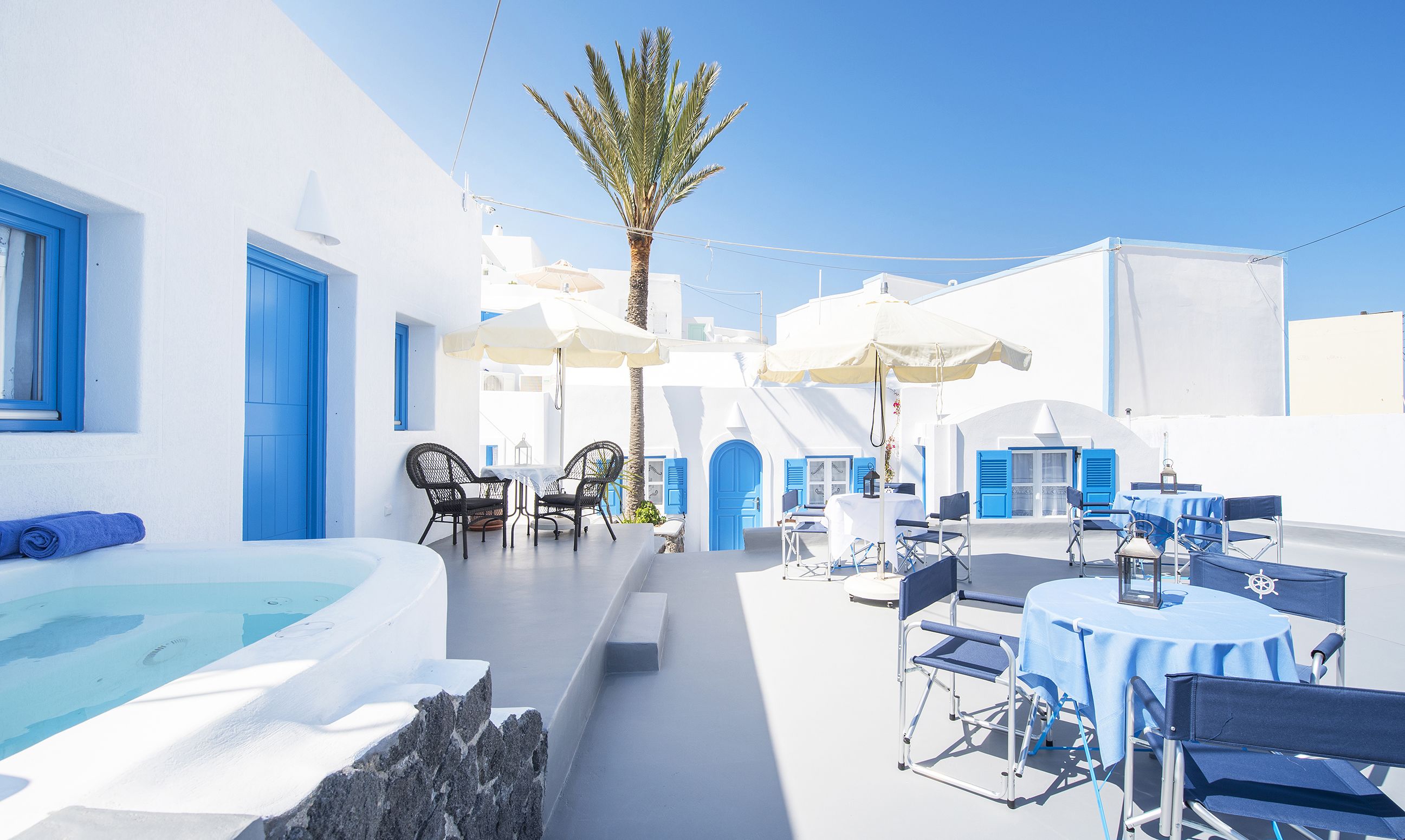 Un espacio al aire libre iluminado por el sol, con piscina, puertas azules y edificios blancos.