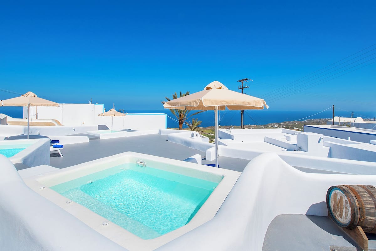 A rooftop pool area with clear blue skies, white architecture, and a view of the sea.