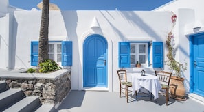 A white building with blue doors and shutters, a stone wall, and a table with chairs outside. - Kadia Traditional Residences (Santorini)