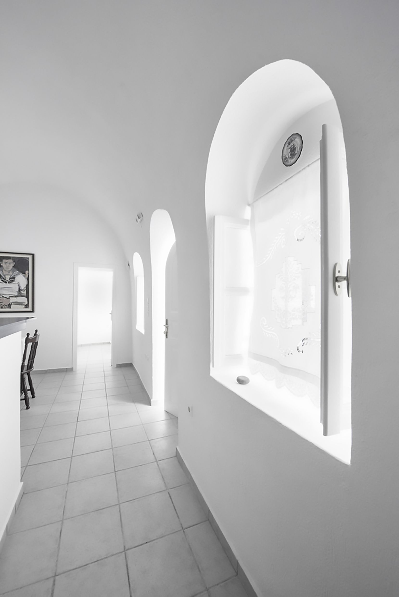 Un pasillo con paredes blancas y suelo de baldosas que conduce a una habitación con un amplio ventanal arqueado y una puerta.