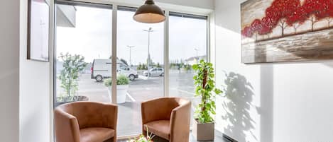 A modern hotel lobby with two leather armchairs, a small table with a vase of flowers, a large painting of red trees, and a view of the parking lot through large windows.