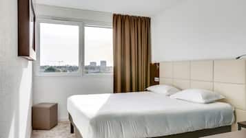Ein Hotelzimmer mit einem großen Bett, einem Nachttisch, einer kleinen Nachttischlampe, einem Fenster mit Vorhängen und Blick auf die Stadt.