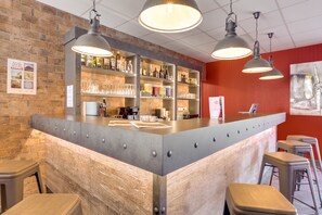A bar area with a wooden counter, metal shelves with bottles, glasses, and a menu. There are stools and a red wall with a painting. - The Originals City, Hôtel Caen Mémorial (Saint-Contest)