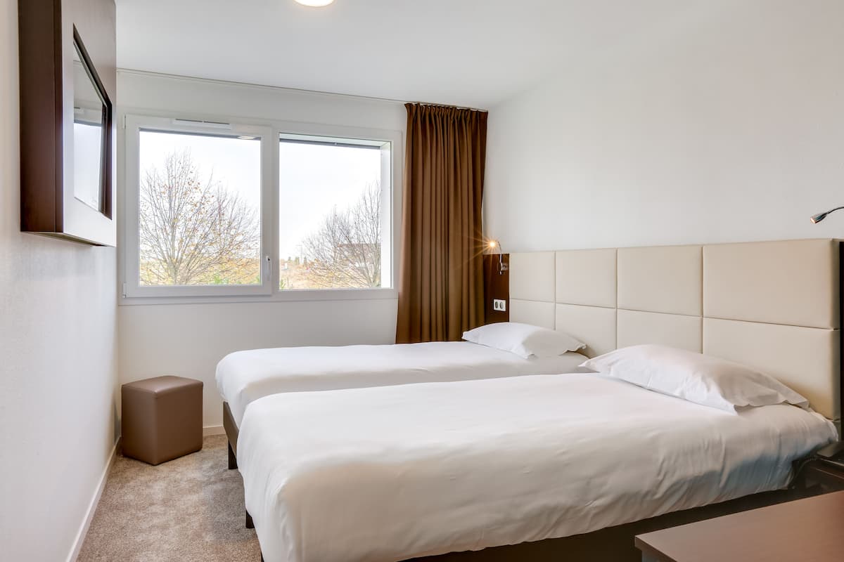 Une chambre d’hôtel avec deux lits, une fenêtre avec des rideaux, une table de chevet et un petit tabouret.