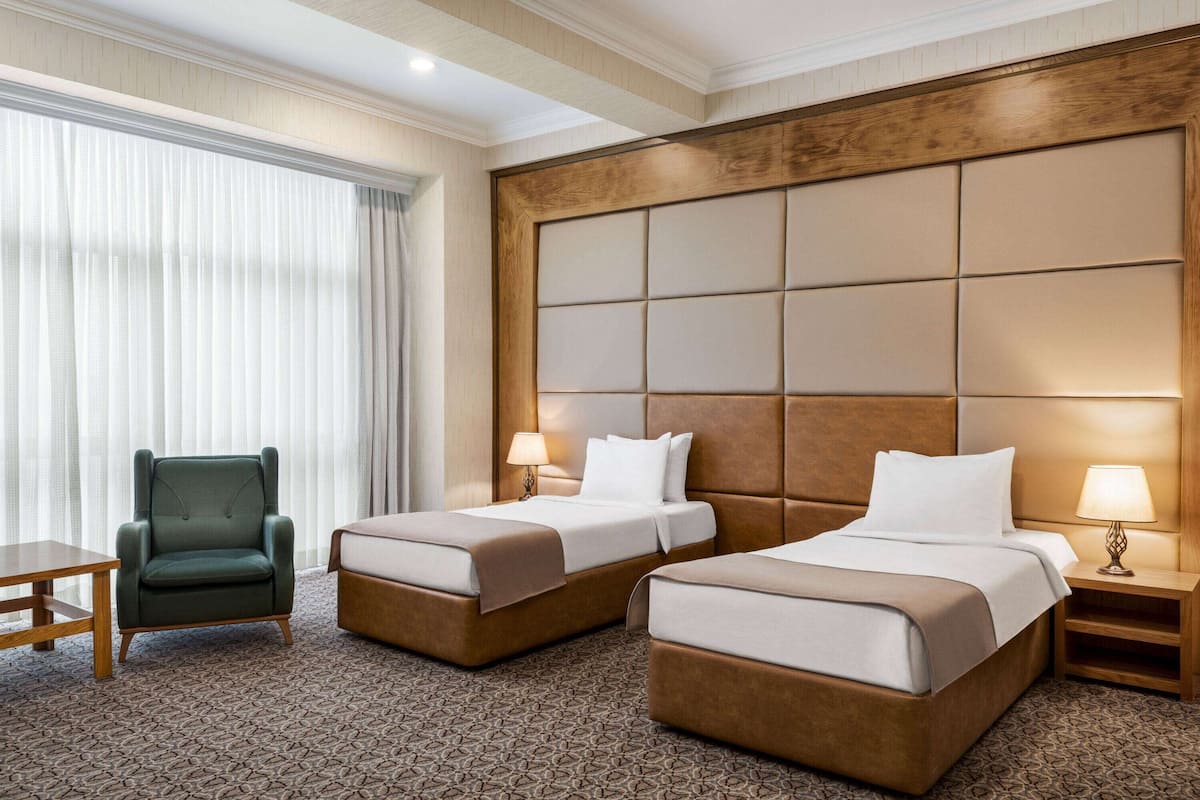 A hotel room with two beds, a green armchair, a wooden nightstand, and a patterned carpet.