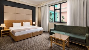 Quarto de hotel com uma cama grande, um sofá verde, uma mesa de madeira e uma janela com cortinas.