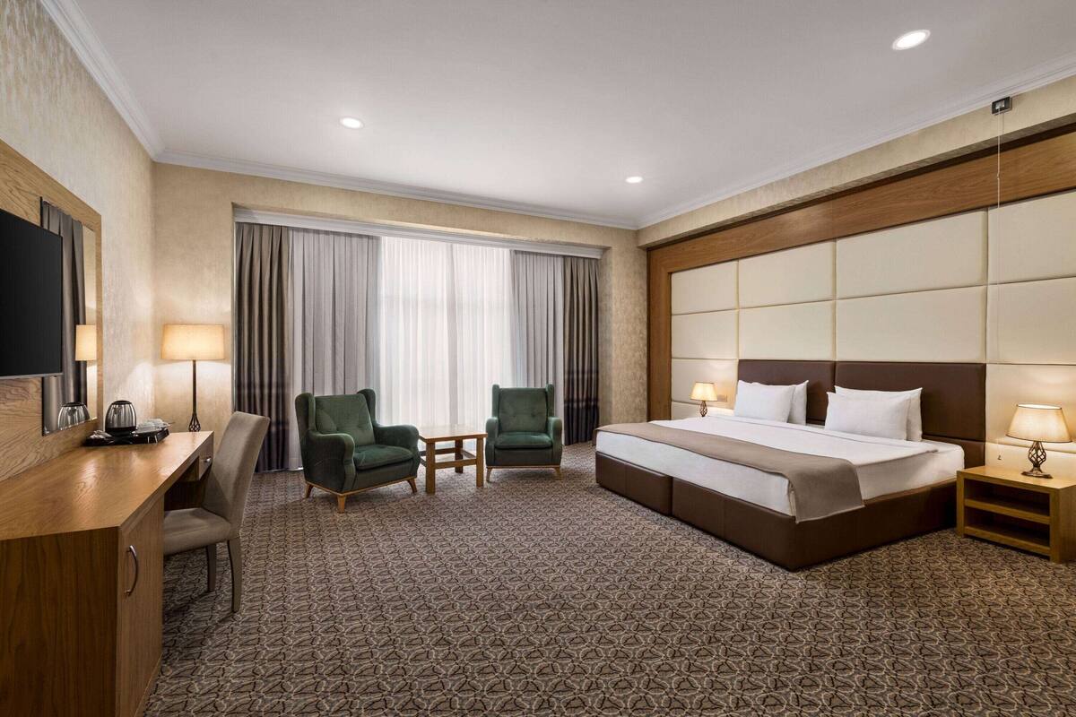 A hotel room with a large bed, two armchairs, a desk with a flat-screen TV, and a minibar.