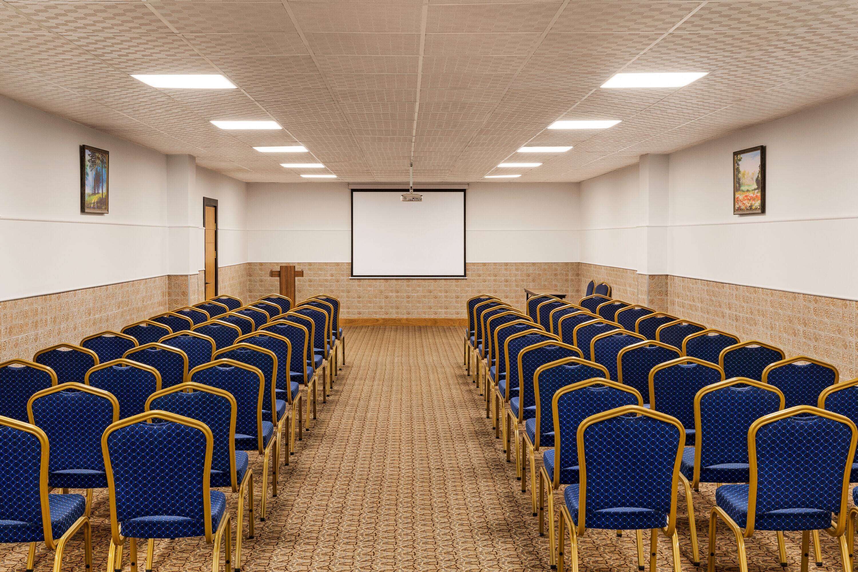 Un salón de conferencias con filas de sillas azules, una pantalla de proyector y suelo alfombrado.