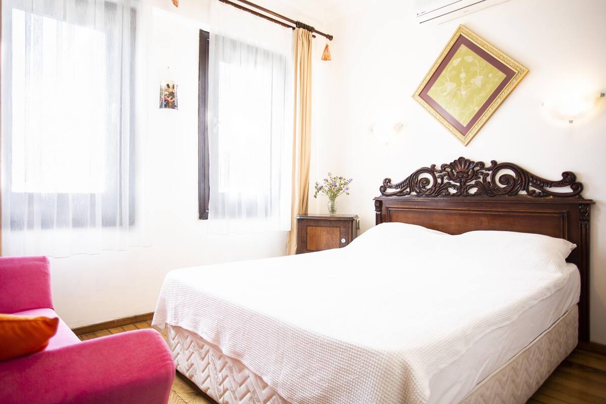 A bedroom with a wooden bed, a pink chair, a nightstand with a vase, and a wall-mounted picture frame.