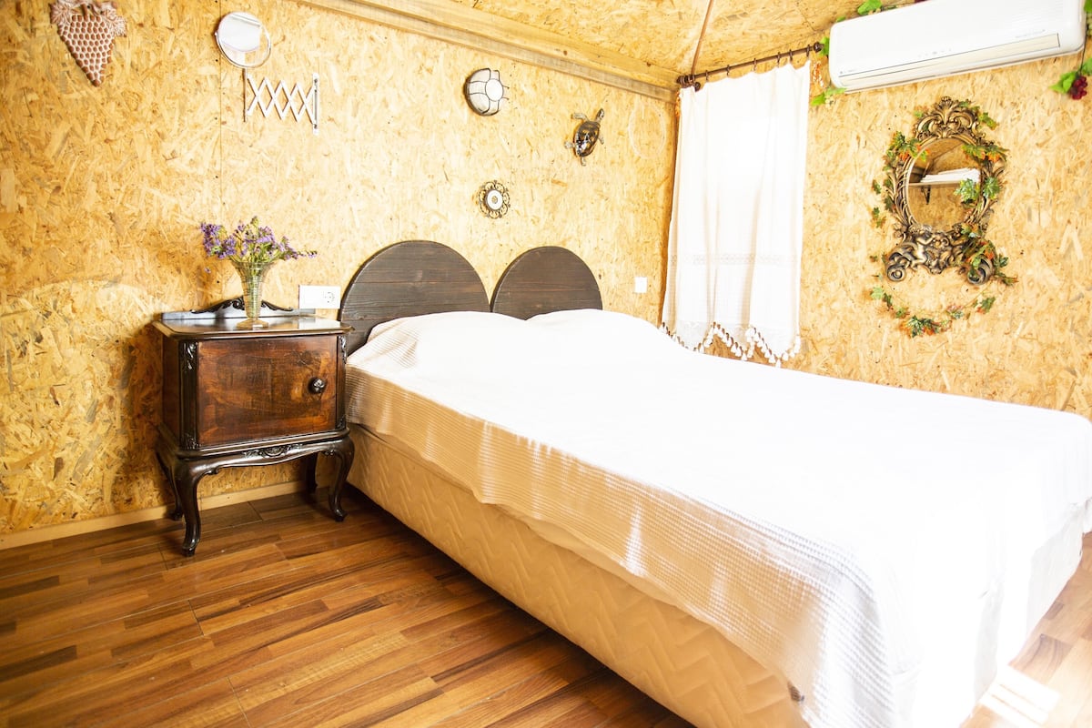 A bedroom with a wooden floor, a bed with a white bedspread, a nightstand with a vase of flowers, and a wall-mounted mirror.