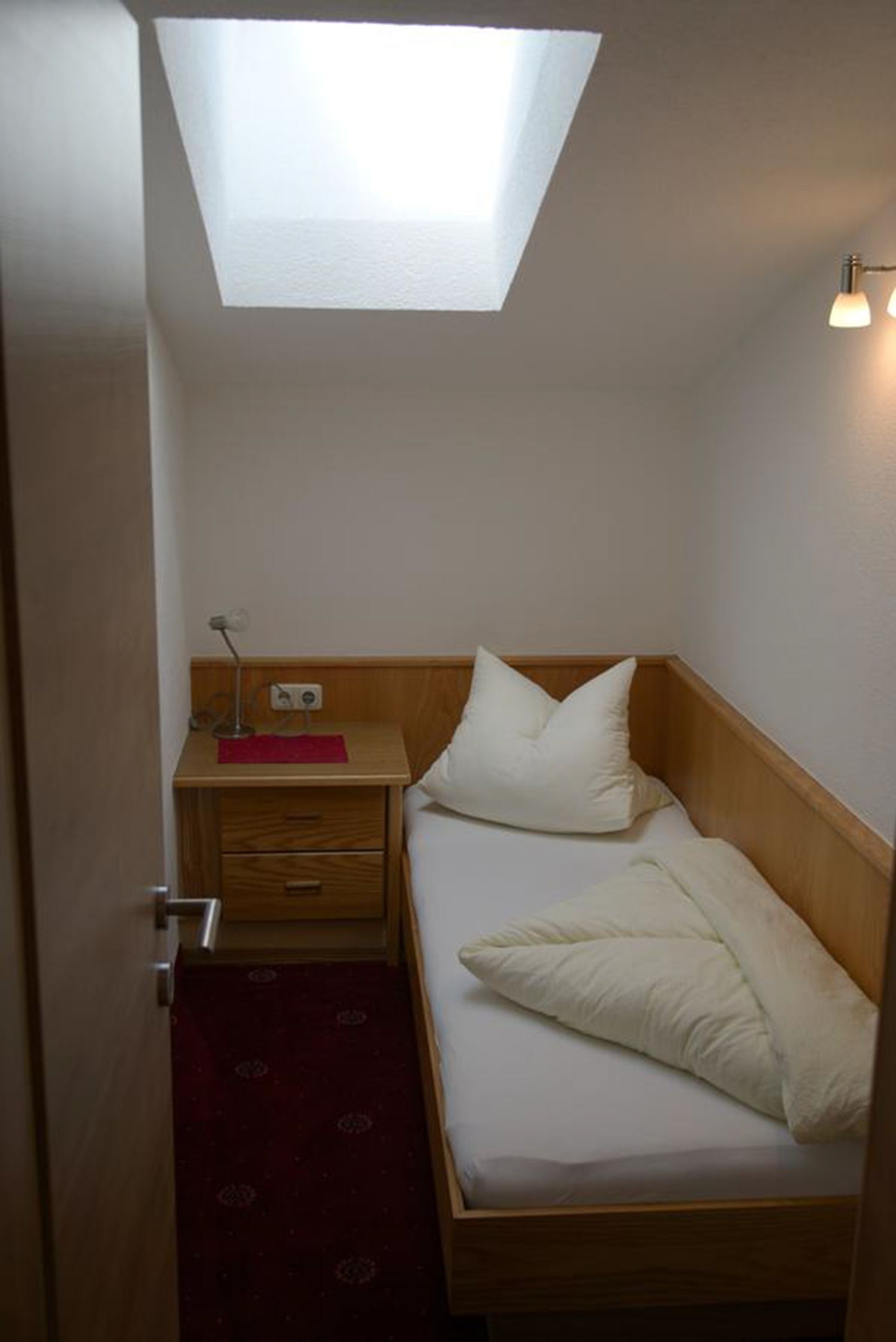 Een kleine eenpersoonskamer met een dakraam, een nachtkastje met lamp en een rood tapijt.