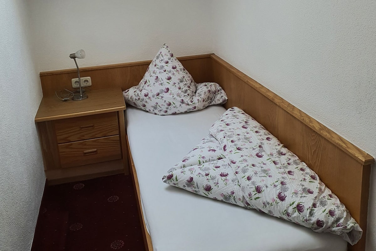 A corner bedroom with a wooden wardrobe, a white bed with floral bedding, and a wall-mounted light fixture.