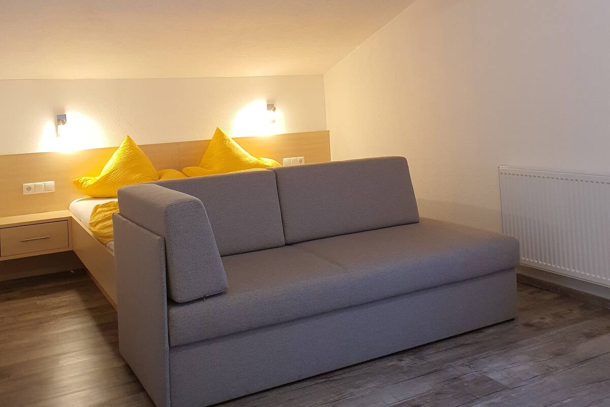 A modern, well-lit bedroom with a grey sofa, a wooden floor, and a bed with yellow pillows.