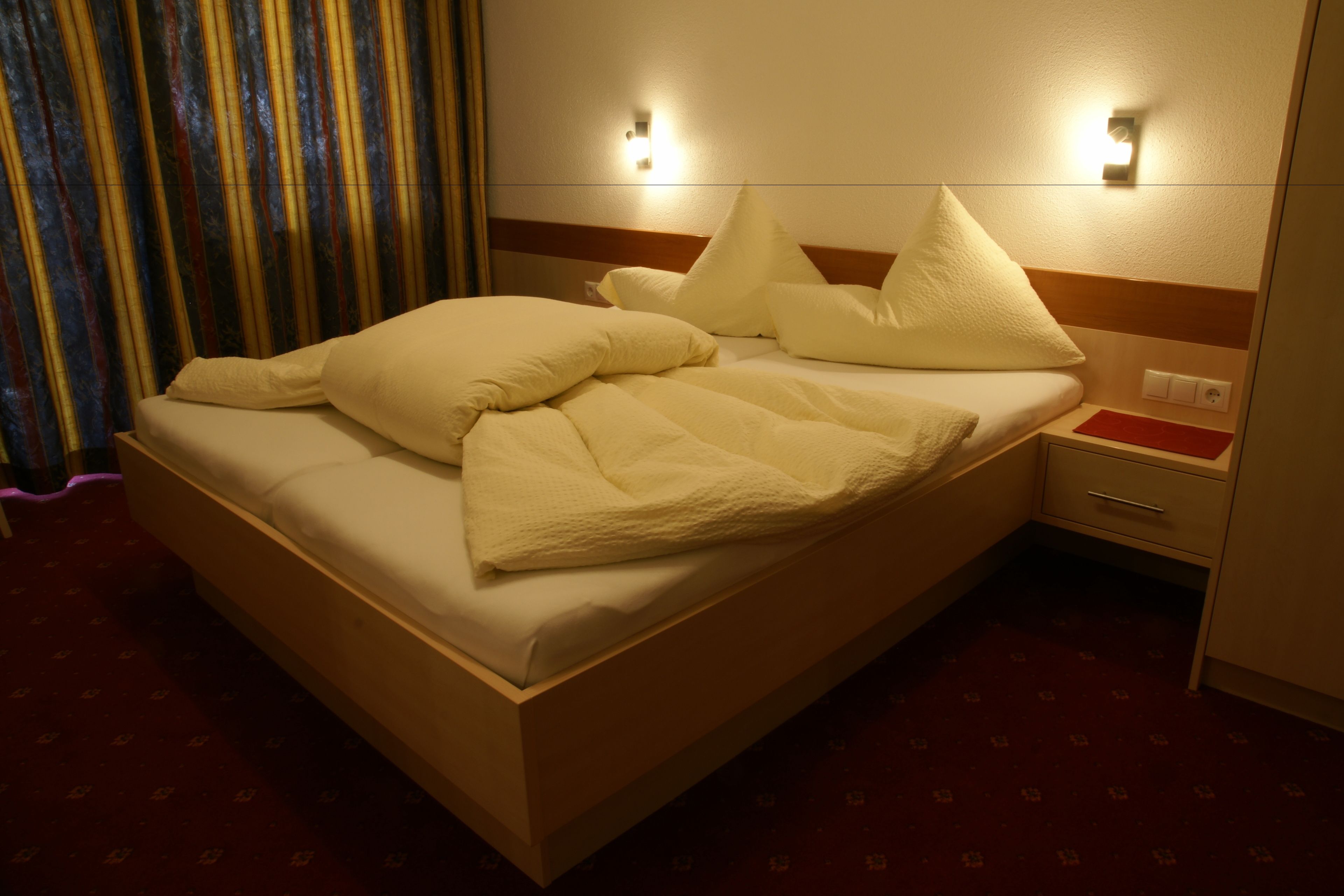 A hotel room with a bed, a nightstand, a red tray table, and a wall-mounted light fixture.