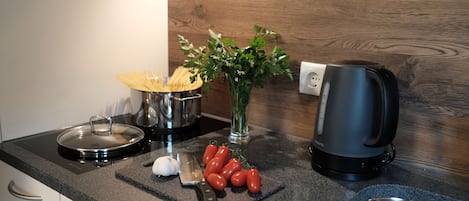 Una encimera de cocina con estufa, hervidor, tabla de cortar con tomates, cuchillo y un florero con hojas verdes.
