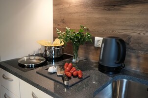 A kitchen counter with a stove, a kettle, a cutting board with tomatoes, a knife, and a vase with green leaves.