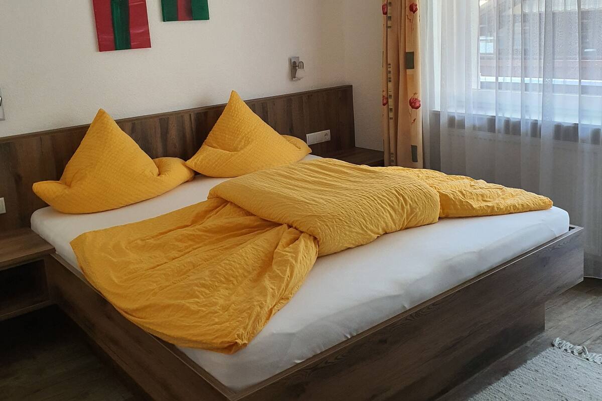A bedroom with a wooden bed, yellow pillows, and a large window with sheer curtains.