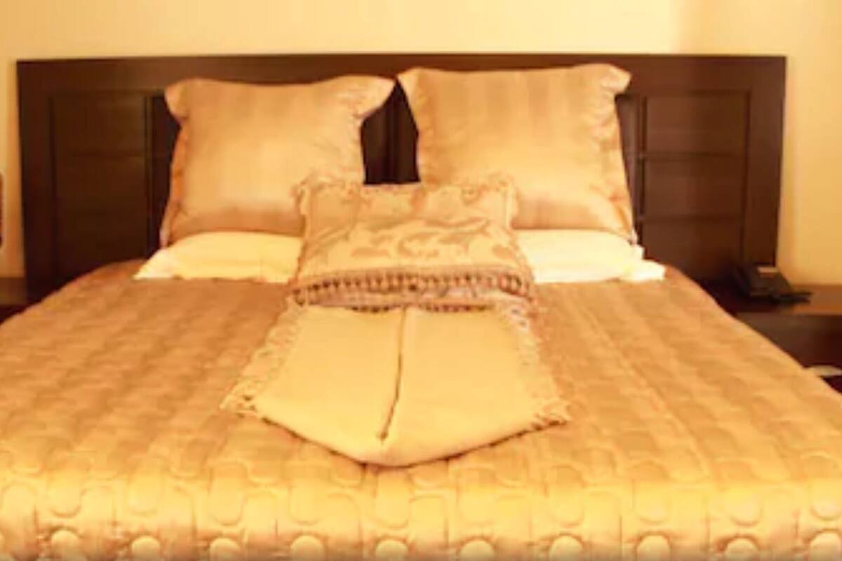 Una cama bien tendida con una colcha estampada, dos almohadas y un cojín decorativo sobre un cabecero de madera oscura.