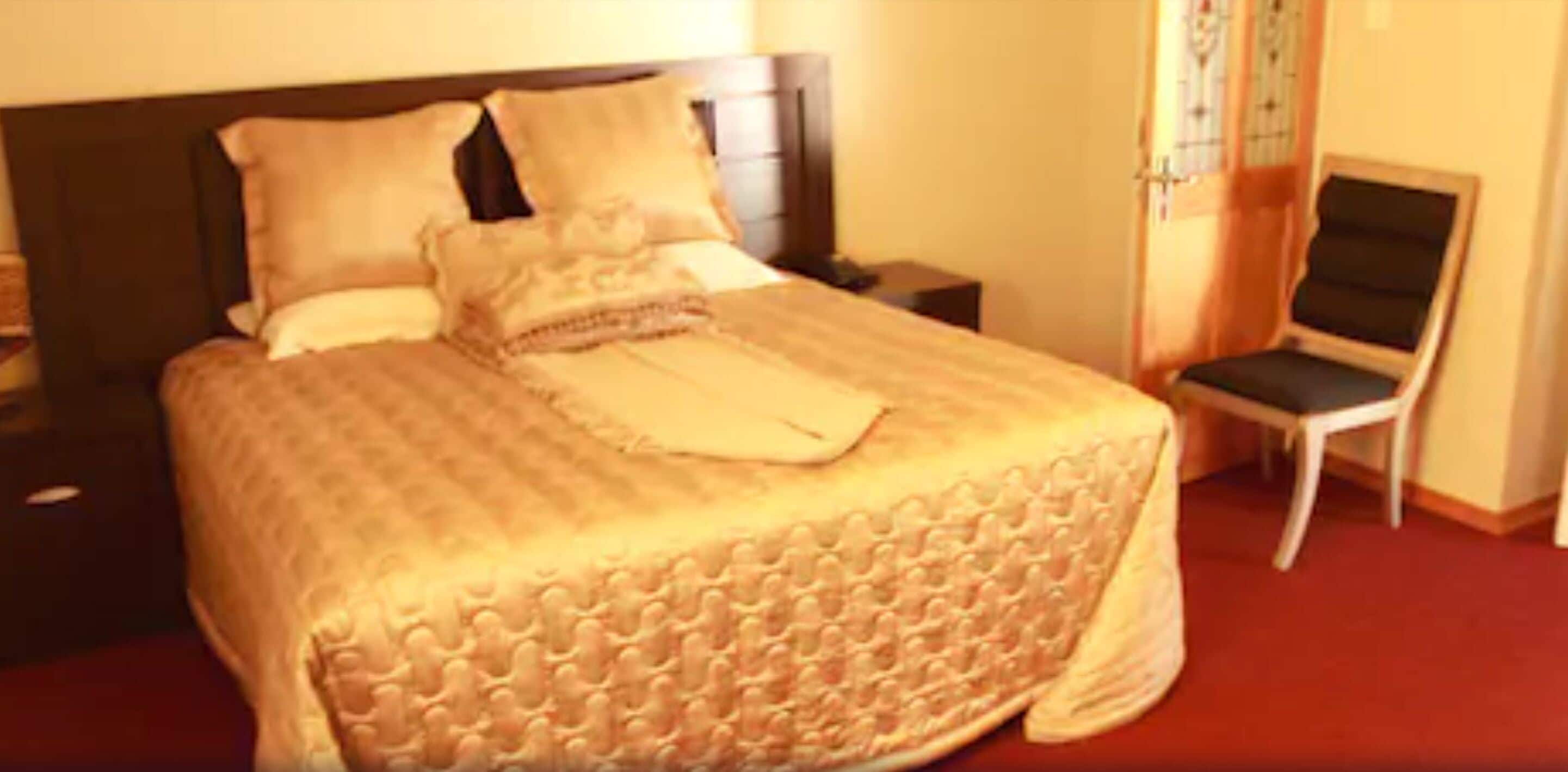 A neatly made bed with a patterned bedspread and pillows, a bedside table with a lamp, and a chair with a cushioned seat.