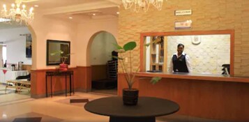 Un área de recepción con recepcionista, un mostrador, una araña de cristal, una planta en maceta y un cartel que dice "WELCOME".