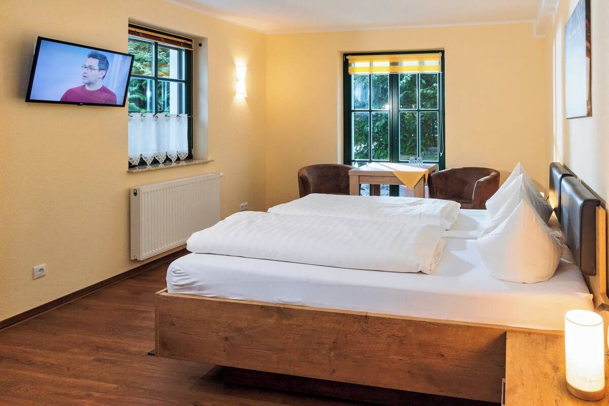 A hotel room with a wooden bed, a television mounted on the wall, a window with curtains, and a table with chairs.